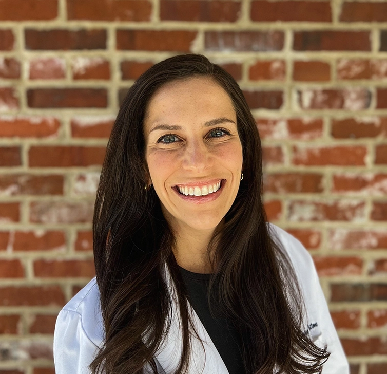 Smiling woman in a white coat against a brick wall backdrop.