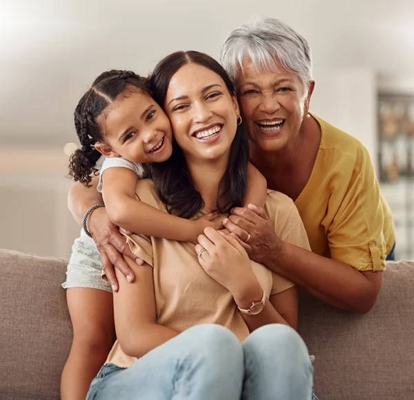 Three generations of women smiling, embracing on a sofa.